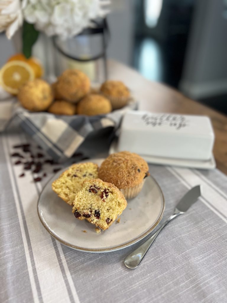 Two cranberry orange muffins on a small plate, one is split open to reveal cranberry bits inside. In the background, there is a basket of muffins, sliced oranges, and dried cranberries.