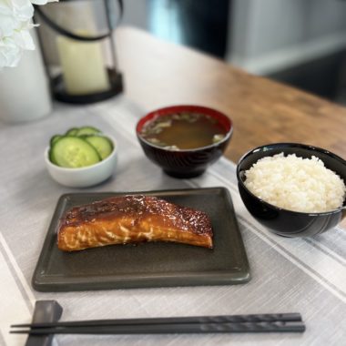 sweet miso salmon on a plate, accompanied by a bowl of steamed short-grain Japanese rice, a bowl of miso soup, and a small plate of salted cucumber pickles