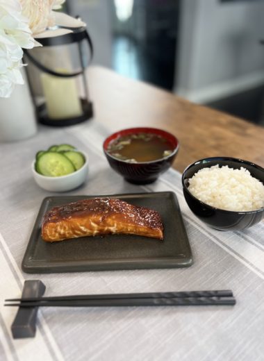sweet miso salmon on a plate, accompanied by a bowl of steamed short-grain Japanese rice, a bowl of miso soup, and a small plate of salted cucumber pickles