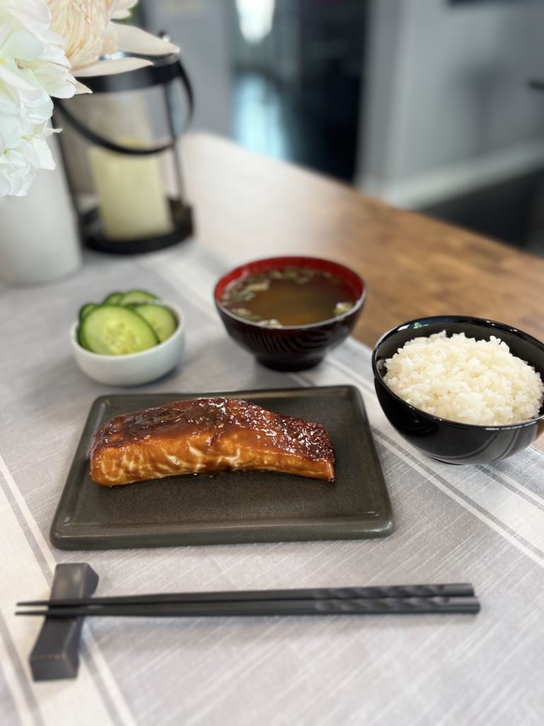 sweet miso glazed salmon on a plate, accompanied by a bowl of steamed short-grain Japanese rice, a bowl of miso soup, and a small plate of salted cucumber pickles