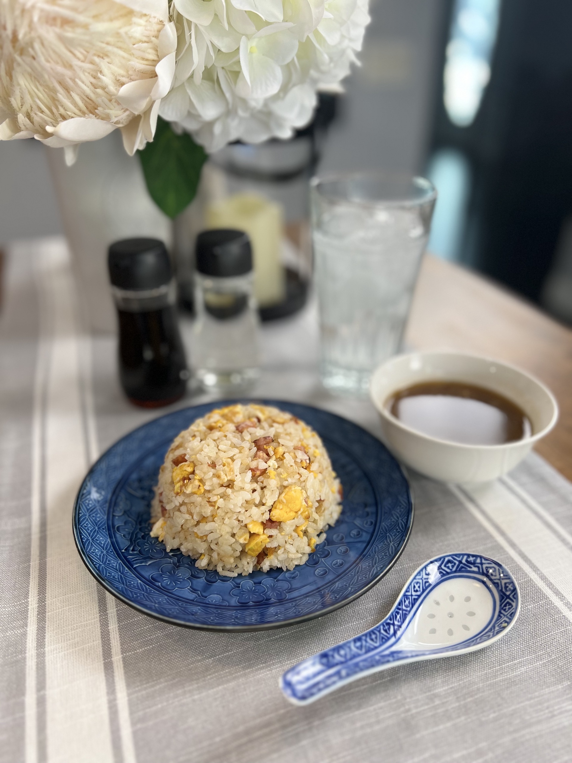 Blue dinner plate with a rounded mound of Japanese style fried rice, known as chahan. Small bottles of soy sauce and white vinegar, a small bowl of pork bouillon soup, and a glass of ice water, in the background. In the foreground, a white and blue ceramic Chinese spoon.